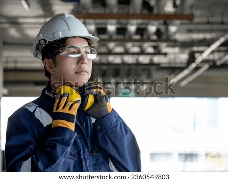 Asian man construction worker wearing uniform suit, safety helmet, goggles and protective gloves holding yellow ear muffs or ear defenders on his neck at construction site