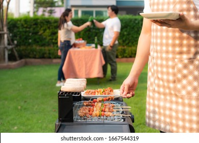 Asian man cooking barbeque grill and sausage for a group of friends to eat party in garden at home. group of friends having outdoor garden barbecue laughing with alcoholic beer drinks - Shutterstock ID 1667544319