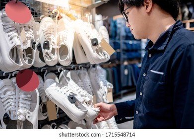 Asian Man Customer Choosing Pair Of White Sneakers In Clothing Store. Buying Stylish Shoes In Fashion Shop. Footwear Industry And Business Concept