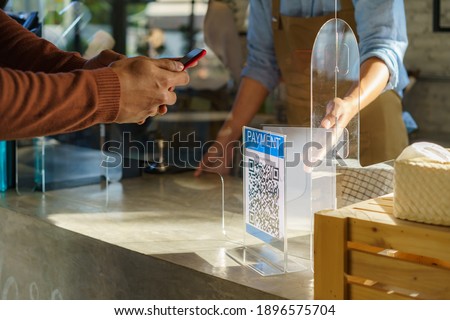 Asian man customer using mobile scan QR or NFC payment on smart phone paying for her coffee drink in coffee shop and plexi screen and Barista wearing mask in background