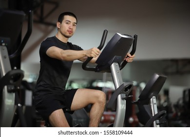 Asian Man Cycling On Bike In Fitness Gym.