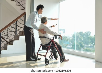 Asian Man Doctor Taking Care Of Elderly Male Patient While Sitting On Wheelchair In A Medical Room Of The Hospital. Health Care, Paralysis, Caregiver.