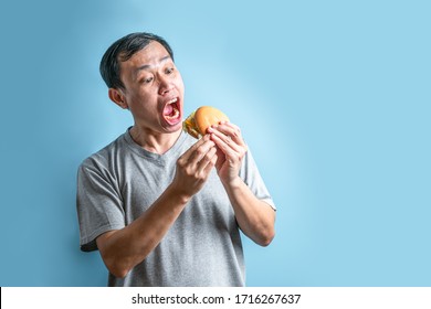 Asian Man Eating Tasty Burger On Isolated Blue Background. Casual T-shirt Lifestyle Portrait