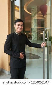 Asian Man In Elegant Black Suit Opening Glass Doorway Of Hotel Greeting Guests 