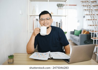 Asian man enjoy drinking coffee while working with paper documents and using laptop at home office. Remote job concept. - Powered by Shutterstock