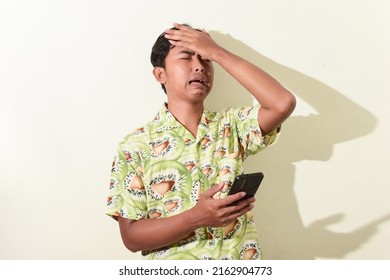 Asian Man Expression Regret While Holding Smartphone And Smacking Forehead. Indonesian Man In Hawaiian Shirt On White Background Isolated. Asian Man Expression Sad Looking At Smartphone Screen