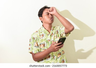 Asian Man Expression Regret While Holding Smartphone And Smacking Forehead. Indonesian Man In Hawaiian Shirt On White Background Isolated. Asian Man Expression Sad Looking At Smartphone Screen