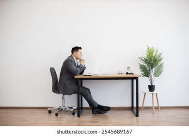 Asian man in formal suit sitting at modern desk working with laptop. White wall background and minimal decor create calm office setting. Businessman appears focused and contemplative. - Powered by Shutterstock