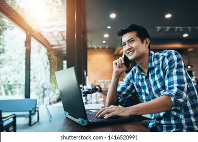 Asian man freelance work on computer touch pad while talking on smart phone With a happy smile, young business man use laptop sitting at wooden table of modern coffee shop, freelance man. - Powered by Shutterstock