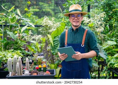 Asian Man Gardener Caring Plants In The Garden. Happy Male Plant Shop Owner Working On Digital Tablet For Checking Plants Order In Store. Small Business Entrepreneur Working With Technology Concept.