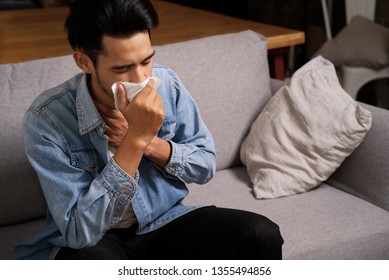 Asian Man Getting A Cold Or Flu Sitting On Sofa At Home. He Is Coughing To Clear The Throat With Hand Holding Tissue Paper Closing His Mouth. Seen From Top Side View. Illness And Sickness Concept.