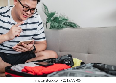 Asian Man Glasses Is Preparing Clothes In Suitcases. He's Choosing Clothes, Travel Documents, The Itinerary For A Solo Trip, And Check The Checklist. Travel, Holiday And Vacation Concept.