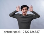 An Asian man in a gray long-sleeve shirt making a silly face, sticking out his tongue while holding both hands up to his face with palms open. The background is white