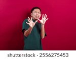 Asian man in a green T-shirt making a scared or surprised expression with his hands raised in defense against a vibrant red background, emphasizing emotions of fear, shock, or reaction