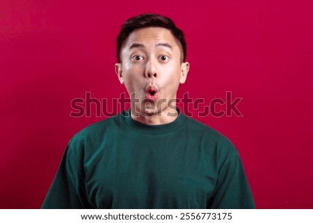 Asian man in a green T-shirt with a surprised and exaggerated expression, hands raised near his face, against a vibrant red background, conveying shock, amazement, or excitement in a playful way