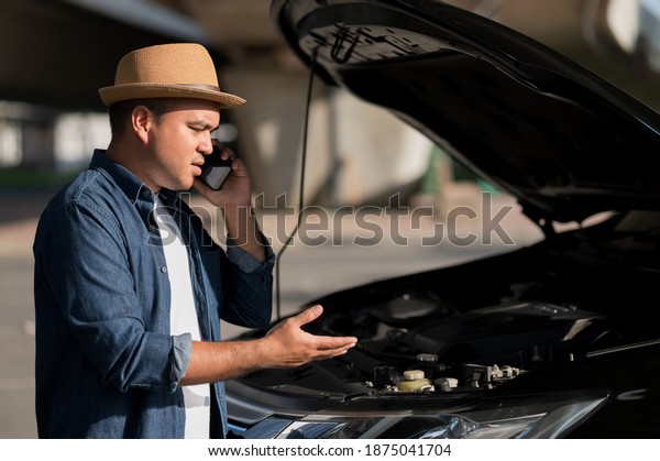 Asian man has  broken down car on the road he\
talking on phone to call someone to help or mechanics car service.\
Emergency assistance\
service