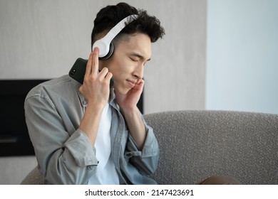 Asian Man In Headphones Listen To Dynamic Music Shaking Head Sitting On Sofa. Young Guy In Casual Clothes Enjoys Weekend Holding Phone At Home