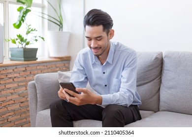 Asian Man Holding Mobile Phone And Looking At It With Smile While Sitting On The Couch At Home.