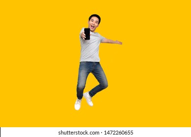 Asian Man Jumping And Presenting Mobile Phone Isolated On Yellow Background