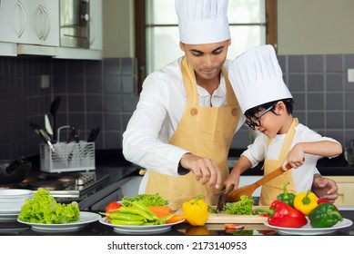 Asian Man With Kid In Chef Hat Apron Helping  Prepare Cooking Fresh Salad Food In Kitchen At Home.Cute Little Boy With Handsome Dad Cooking Together.Family Healthy Lifestyle Concept