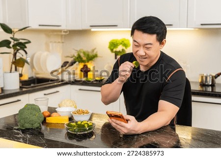 Asian man in kitchen eats broccoli snack and checks health news on orange smartphone. Adult middle-aged man enjoys low calorie snack indoors feels motivated to follow diet plan and lifestyle change.