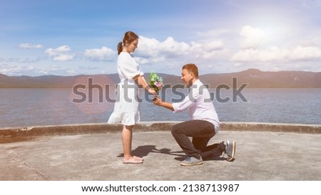 An Asian man knelt down to give a bouquet of flowers to an Asian woman wearing white at the sea port. To express love and to propose in the sunshine, clear skies, strong winds.