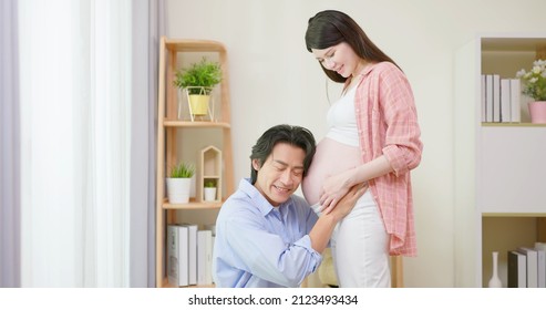 Asian Man Is Listening Pregnant Wife Belly And Expecting Their Baby Coming Near Window At Home