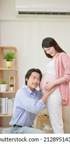 Asian Man Is Listening Pregnant Wife Belly And Expecting Their Baby Coming Near Window At Home