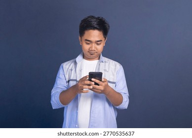 Asian man looking down and using his smartphone with a neutral expression on a blue background.