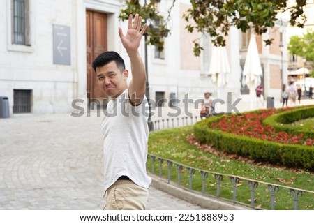 Asian man looking sad while waving hand to say goodbye to someone outdoors in the street.