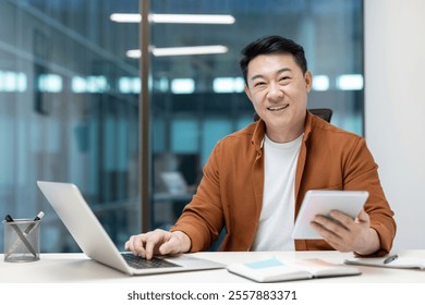 Asian man mature businessman working in an office using a tablet and laptop, smiling confidently. The atmosphere reflects productivity, success, and a positive attitude. - Powered by Shutterstock