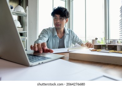 Asian Man Messy Hair Working In Architect Or Interior Designer Study About Building Design With Laptop And Drawing Paper, Building Model On Desk In Bright Home Office. Technology For Design Freelance