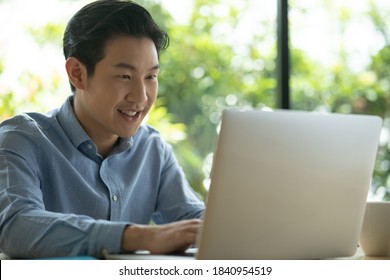 Asian Man On Blue Shirt Smiling While Using A Laptop. Happy Face Smiling Asian Short Hair Man Looking At A Laptop With Green Background. Freelancer Working And Looking At Laptop. 