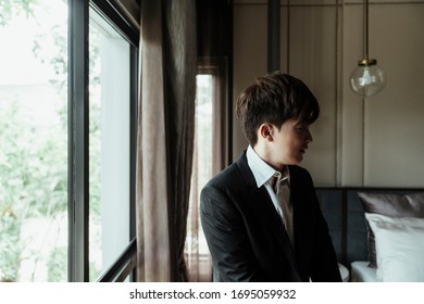 Asian Man On White Collar Shirt Dressing Up Black Suit In Bedroom Near Window.