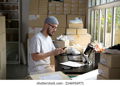 Asian Man Online Seller Confirming Orders From Customer On The Phone. E-commerce Male Business Owner Looking At The Phone In Store Warehouse.