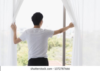 Asian man opening the curtain and looking outside the window.