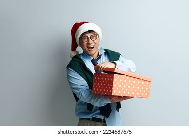 Asian Man Opening Present. Happy Excited Guy In Santa Hat Holding In Hands Half Opened Red Wrapped Present Box, Looking Camera With Excitement And Surprise. Indoor Studio Shot Grey Background 