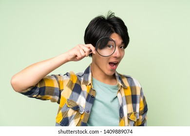 Asian Man Over Isolated Green Wall Holding A Magnifying Glass