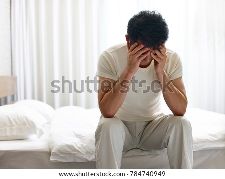 asian man painful and miserable sitting on bed head down covering face with hands.