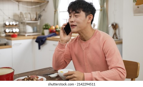 Asian Man Picking Up Call While Eating Lunch Alone At Home. He Puts Down Chopsticks And Ball To Chat On The Phone With Friends In Dining Room