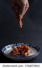 An Asian Man Picks Up Fried Chicken Skin From A Thai Patterned Dish On A Black Background.