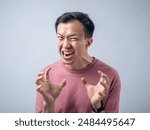 An Asian man in a pink shirt exhibits a fierce expression, baring his teeth and raising his hands with claw-like gestures, as if in anger or frustration. The plain light blue background highlights