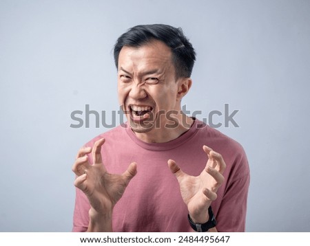 An Asian man in a pink shirt exhibits a fierce expression, baring his teeth and raising his hands with claw-like gestures, as if in anger or frustration. The plain light blue background highlights