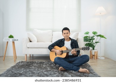 Asian man playing guitar sitting on sofa couch or floor in the living room at home. Asian man writing song while playing guitar at home. Compose song music concept.