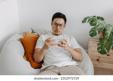 Asian man playing mobile game making funny intense face in the living room.