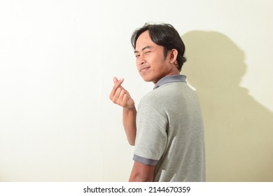 Asian Man Poses Smiling And Crossing His Index Finger And Thumb. Indonesian Man In Gray T-shirt On White Background Isolated. The Symbol Of Sarangheo, Saranghae, Saranghaeyo. Korean Teen Pose