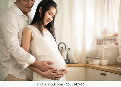 Asian Man And Pregnancy Woman Stand In Kitchen At Home. Father Holding Mother Tummy Feeling Love Expecting About Future Newborn Baby. Husband Hugs Wife From Behind. Love And Care Of Pregnant Concept.