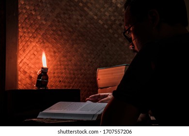 Asian Man Reading Book On An Old Wood Table In A Rustic House Room Lit By A Traditional Oil Lamp From A Used Medicine Bottle. The Concept Of Education In Countrysides Or Work Or Study From Home