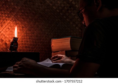 Asian Man Reading Book On An Old Wood Table In A Rustic House Room Lit By A Traditional Oil Lamp From A Used Medicine Bottle. The Concept Of Education In Countrysides Or Work Or Study From Home