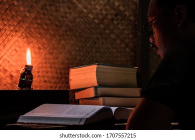 Asian Man Reading Book On An Old Wood Table In A Rustic House Room Lit By A Traditional Oil Lamp From A Used Medicine Bottle. The Concept Of Education In Countrysides Or Work Or Study From Home